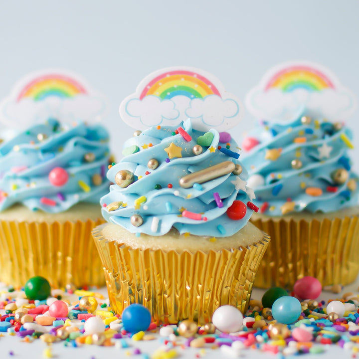 Three decorated cupcakes with light blue frosting, gold wrappers, rainbow toppers, and colorful sprinkles on a white surface with scattered sprinkles.