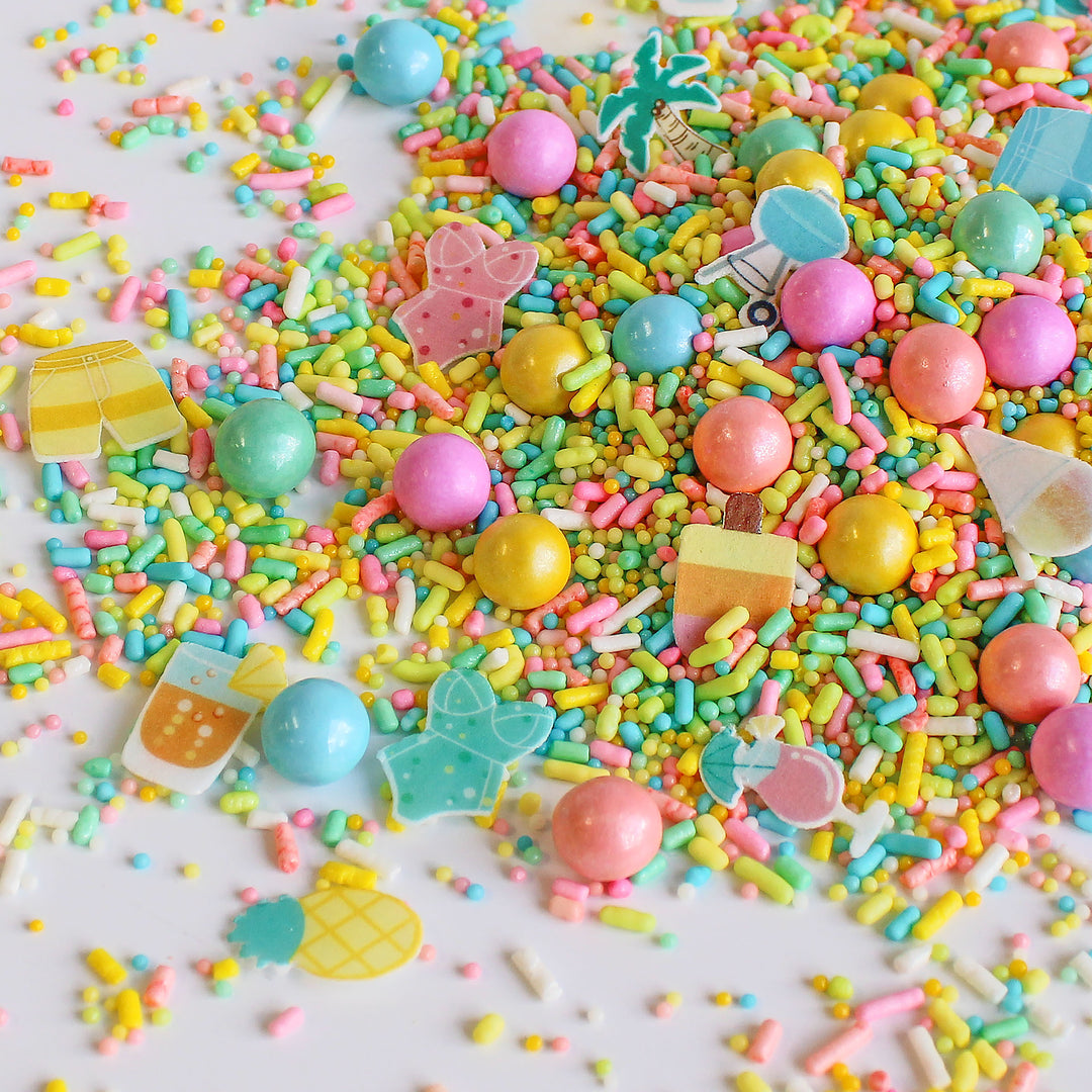 A close-up photo of Palm Springs Sprinkle Mix with a variety of colorful sprinkles, including specialty pieces like wafer swimsuits, palm trees, snow cones, umbrella drinks, and bbq grills. Perfect for decorating cakes, cookies, cupcakes, and even ice cream!