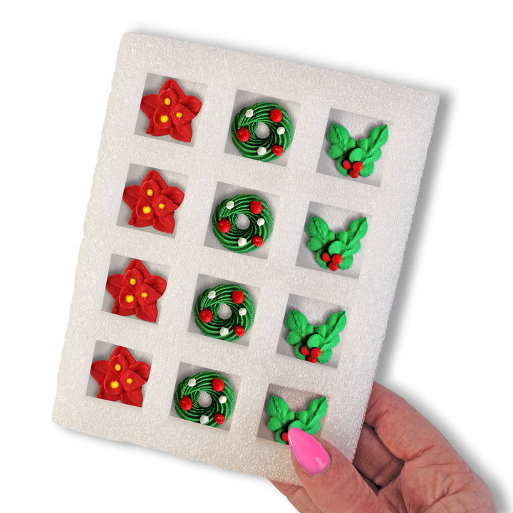 A hand holding a tray of 12 royal icing holiday decorations, featuring red poinsettias, green wreaths, and holly, against a white background.