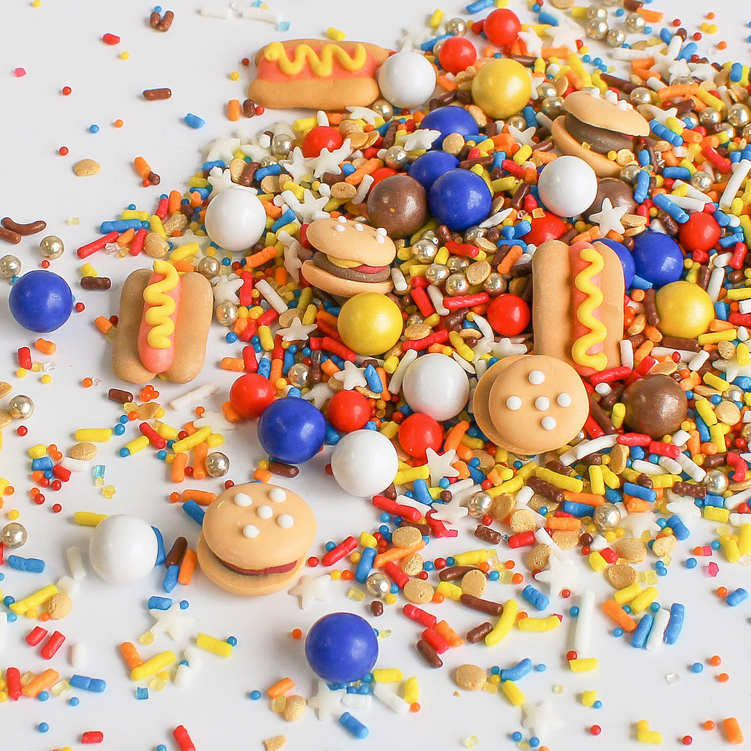 A photo of the Summer BBQ Sprinkle Mix featuring red, blue, and yellow sprinkles as well as royal icing hamburgers and hot dogs.