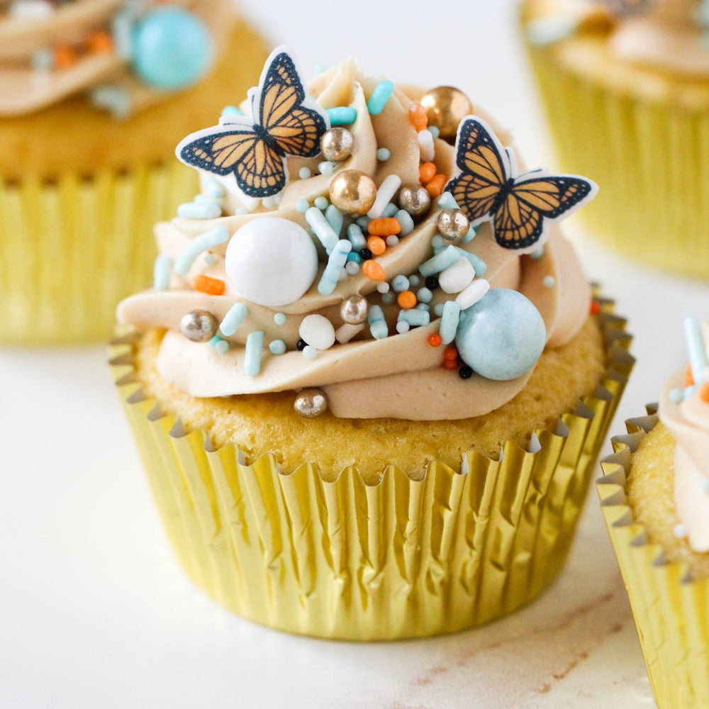 A cupcake with Monarch Butterfly Sprinkle Mix decorations, featuring light blue sprinkles and black and orange butterfly wafer paper decorations.