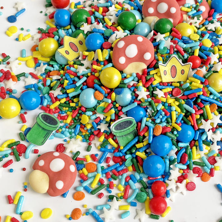 A close-up photo of the "It's Me" Sprinkle Mix in a resealable container. The mix features red, blue, green and yellow sprinkles with royal icing mushroom caps and wafer paper green pipes and princess crowns. Use them to decorate your favorite video game-themed baked goods for a playful touch.